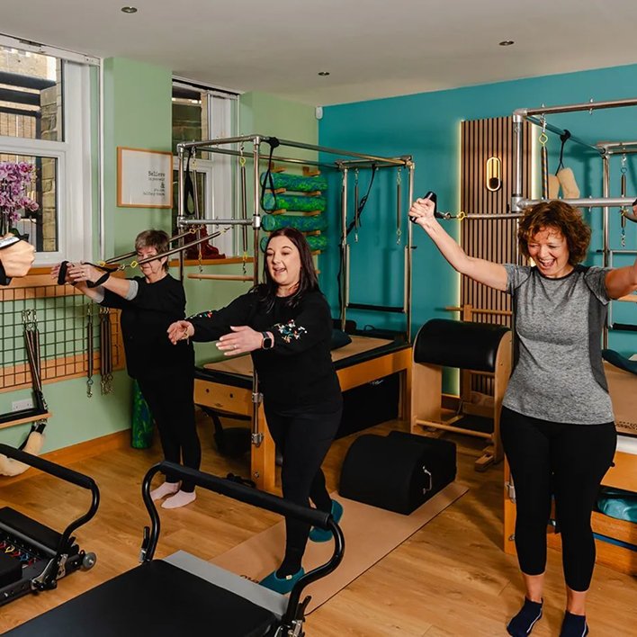 Three smiling women exercising together in a small group class, surrounded by studio equipment