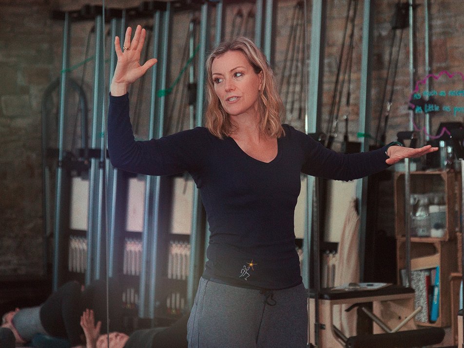 Helen, founder and teacher at The Movement Retreat, leading a class in a rustic brick-walled studio. She is wearing a dark long-sleeve top and grey joggers, with her arms outstretched as she guides her students. Pilates equipment and class participants can be seen in the background.