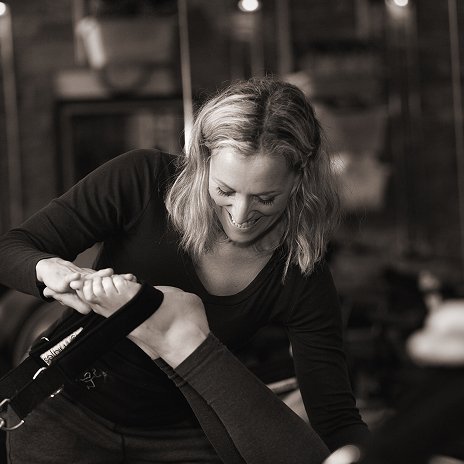 A black and white photo of Helen, founder and teacher at The Movement Retreat, smiling warmly as she provides hands-on guidance to a student during a Pilates session in her studio.