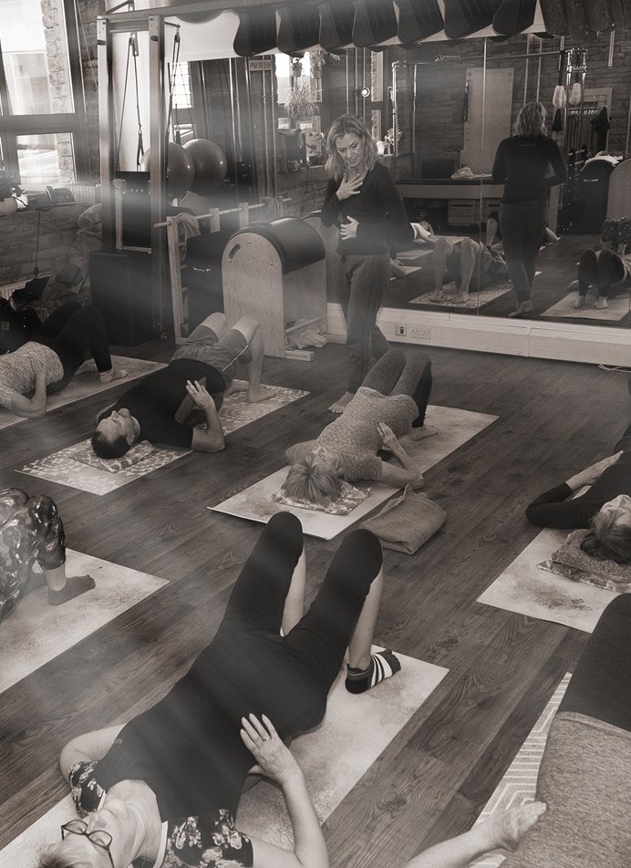 A black and white photo of Helen, founder of The Movement Retreat, teaching a Pilates mat class in her rustic brick-walled studio. Several students lie on mats performing a floor exercise while Helen stands and observes, with Pilates equipment and studio mirrors visible in the background.
